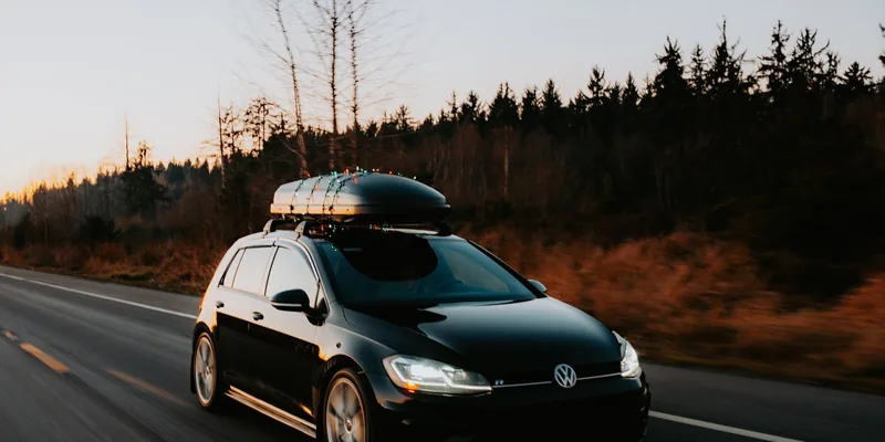 Grote dakkoffer op de dakdragers van een zwarte auto die bij zonsondergang over een bosweg rijdt