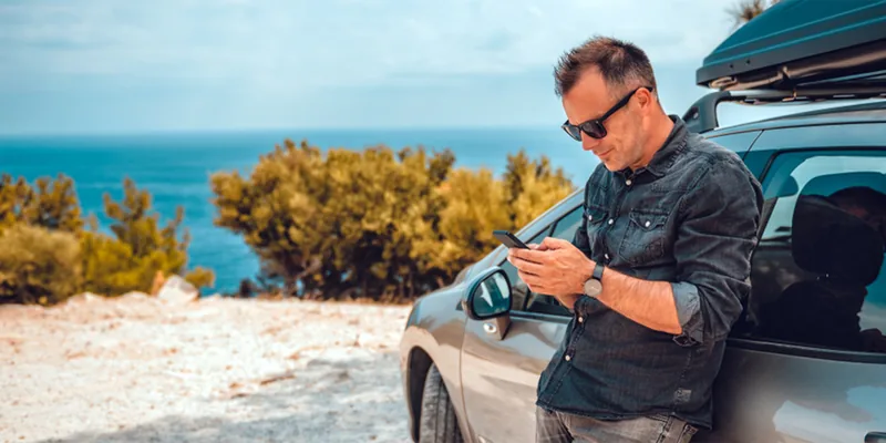 Man kijkt op zijn telefoon naast een auto met dakkoffer aan de kust