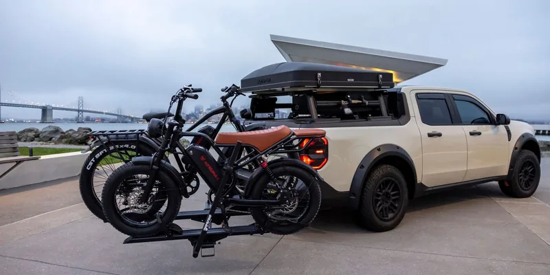 Auto met dakkoffer en twee e-bikes op de fietsendrager bij een brug